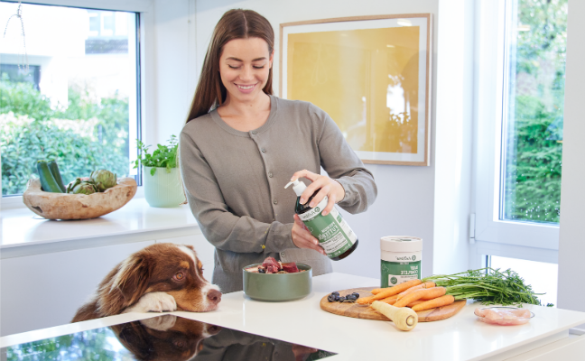 Woman in a kitchen holding a bottle of Anifrote Premium Feed Oil, with a dog and various items on the counter.
